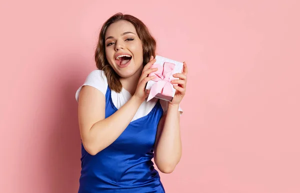 Jovem menina bonita animado com cabelo encaracolado longo segurando pequena caixa de presente isolado no fundo rosa. Conceito de emoções, beleza, moda, feriados — Fotografia de Stock