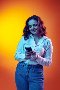Uzun kıvırcık saçlı, turuncu arka planda izole edilmiş akıllı telefon kullanan heyecanlı, genç ve güzel bir kız. Duygular, güzellik, moda kavramı