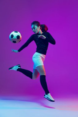 Futbol topuyla antrenman yapan bayan futbolcunun dinamik portresi neon ışıklarıyla izole edilmiş mor stüdyo arka planında. Spor, eylem, hareket, fitness