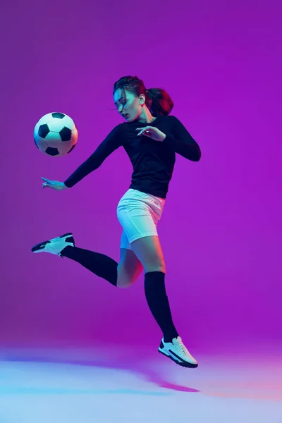 Dynamic portrait of female soccer player practicing with football ball isolated on purple studio background in neon light. Sport, action, motion, fitness — Stock Photo, Image