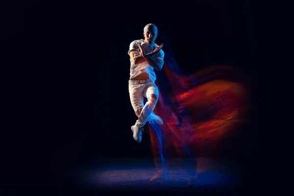 Skákání. Stylový muž ve sportovně bílém obleku tančící hip-hop, breakdance izolovaný na tmavém pozadí ve smíšeném neonovém světle. Mládež kultura, hip-hop, styl a móda, akce. — Stock fotografie