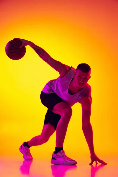 Studio shot van gespierde man, basketbal speler training met bal geïsoleerd op gradiënt geel paarse achtergrond in neon licht. Schoonheid, sport, beweging, activiteitsconcepten. — Stockfoto