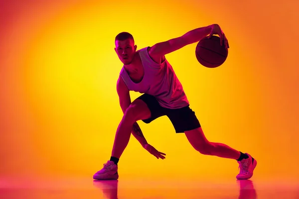 Studio shot van gespierde man, basketbal speler training met bal geïsoleerd op gradiënt geel paarse achtergrond in neon licht. Schoonheid, sport, beweging, activiteitsconcepten. — Stockfoto