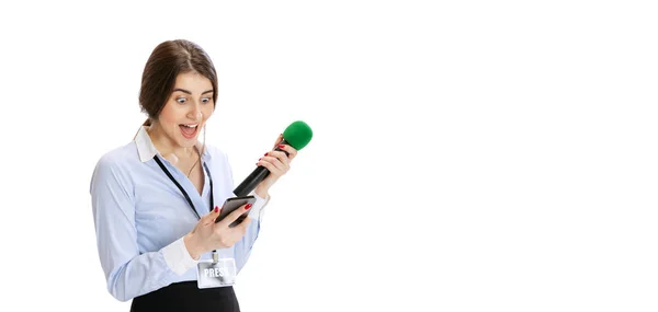Захоплена молода дівчина, жінка-журналістка тримає мікрофон репортера і дивиться на телефон ізольовано на білому тлі. Концепція соціальних мереж, преси, новин, інформації — стокове фото