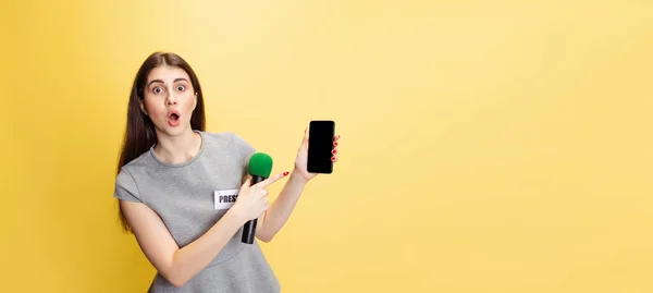 Portrait à mi-longueur d'une jeune fille choquée, correspondante utilisant un téléphone isolé sur fond de studio jaune. Concept de médias sociaux, presse, nouvelles, information — Photo