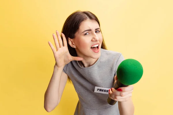 Емоційна молода дівчина, представниця жіночої преси, тримає репортерський мікрофон ізольовано на жовтому студійному фоні. Концепція соціальних мереж, преси, новин, інформації — стокове фото