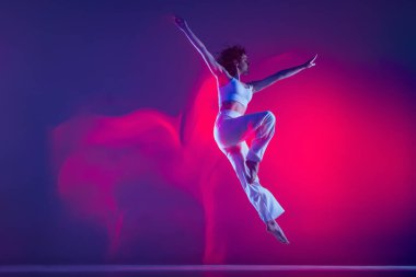 Genç sportif güzel kız, hip-hop dansçısı pembe neon ışıkta mor arka planda izole bir şekilde zıplıyor. Gençlik kültürü, tarz ve moda, aksiyon.