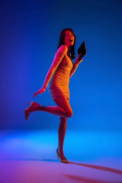 Portret van jong emotioneel meisje, student met lang donker haar met behulp van gadget geïsoleerd op donkerblauwe achtergrond in neon licht. Concept van emoties — Stockfoto