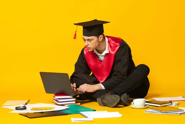 Портрет молодого человека, студента в выпускном колпаке и проект типирования платья на ноутбуке изолирован на желтом фоне студии — стоковое фото