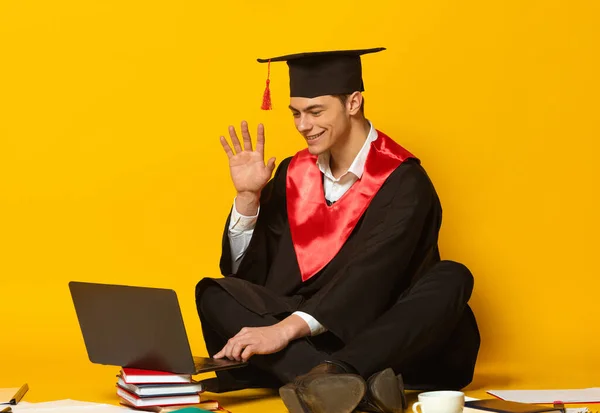 Retrato de un joven, estudiante con gorra de graduación y vestido sentado en el suelo y saludando en el portátil, que tiene lección en línea aislada sobre fondo amarillo —  Fotos de Stock