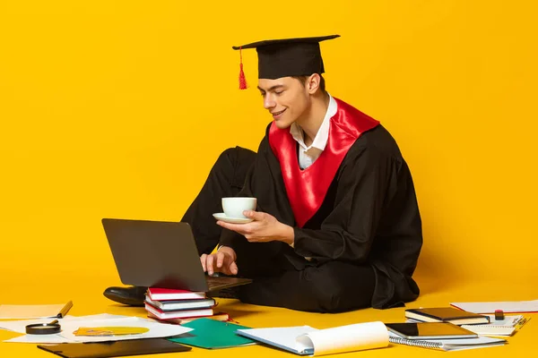 Retrato de hombre joven, estudiante en gorra de graduación y manto sentado frente a la computadora portátil aislado sobre fondo de estudio amarillo —  Fotos de Stock