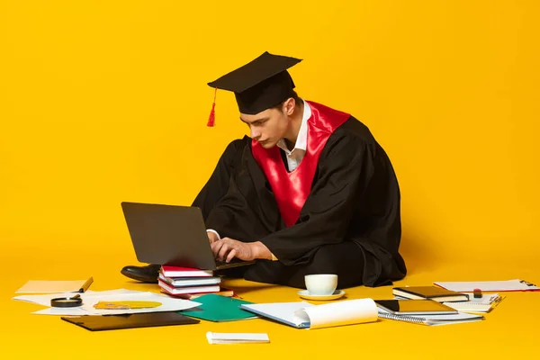 Retrato de un joven, estudiante con gorra de graduación y vestido de trabajo en el ordenador portátil aislado sobre fondo de estudio amarillo —  Fotos de Stock