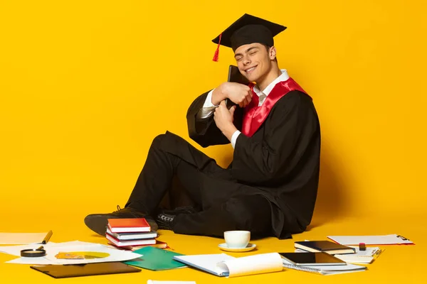 Retrato de hombre joven, estudiante en gorra de graduación y vestido abrazando portátil aislado sobre fondo de estudio amarillo —  Fotos de Stock