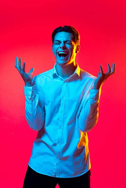 Retrato de jovem de camisa branca gritando e espalhando as mãos isoladas sobre fundo estúdio vermelho em luz de néon — Fotografia de Stock