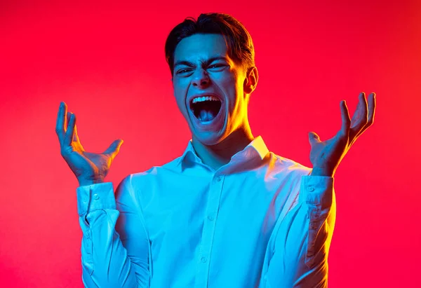 Porträt eines jungen Mannes in weißem Hemd, der schreit und die Hände über rotem Studiohintergrund in blauem Neonlicht ausbreitet — Stockfoto