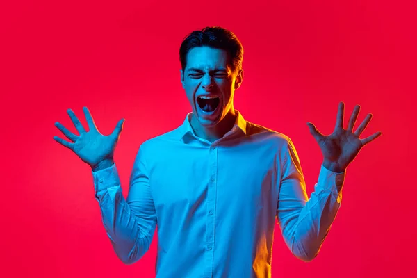 Porträt eines jungen Emotiva-Mannes, Student im weißen Hemd schreiend, die Hände isoliert über rotem Studiohintergrund in blauem Neon ausgebreitet — Stockfoto