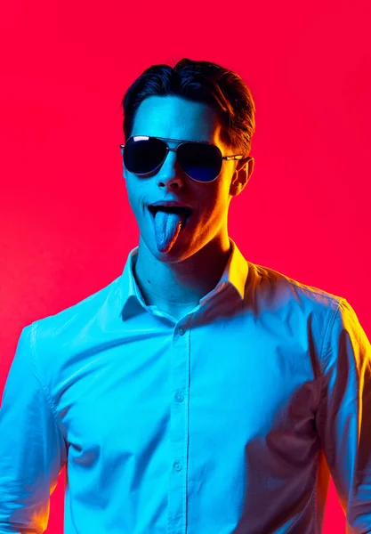 Retrato de homem bonito jovem em óculos de sol elegantes e camisa branca com a língua saliente isolado sobre fundo estúdio vermelho em néon azul — Fotografia de Stock