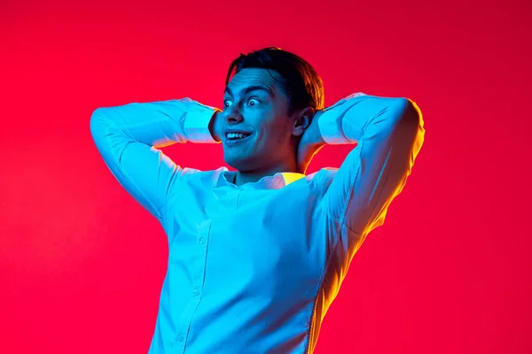 Retrato de jovem homem emotivo, estudante de camisa branca mostrando emoções de emoção isolado sobre fundo estúdio vermelho em néon azul — Fotografia de Stock