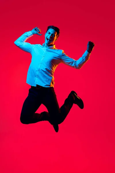 Retrato de jovem homem sorridente, estudante de camisa branca alegremente pulando em excitação isolada sobre o fundo do estúdio vermelho em néon azul — Fotografia de Stock