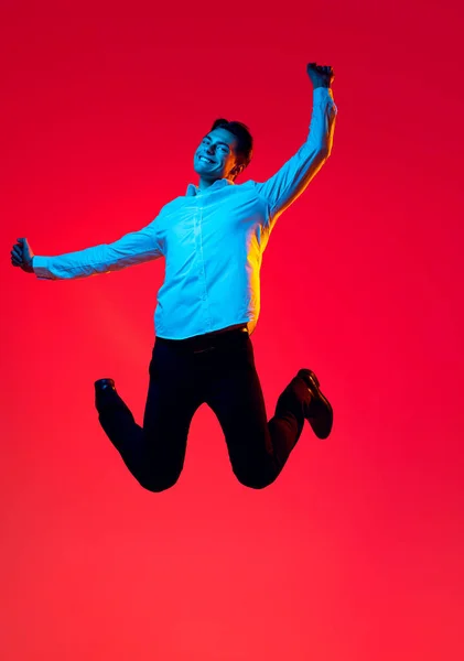 Porträtt av ung leende man, student i vit skjorta glatt hoppa i spänning isolerad över röd studio bakgrund i blå neon — Stockfoto