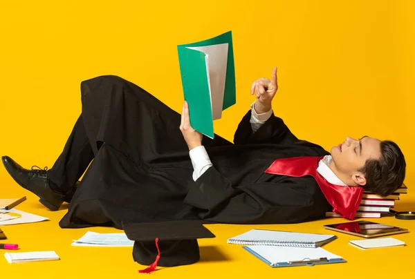 Retrato de hombre joven, estudiante en graduación mostrada tumbado en el suelo y papeles de lectura aislado oer fondo de estudio amarillo —  Fotos de Stock