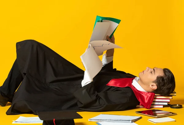 Retrato de hombre joven, estudiante en graduación mostrada tumbado en el suelo y papeles de lectura aislado oer fondo de estudio amarillo —  Fotos de Stock