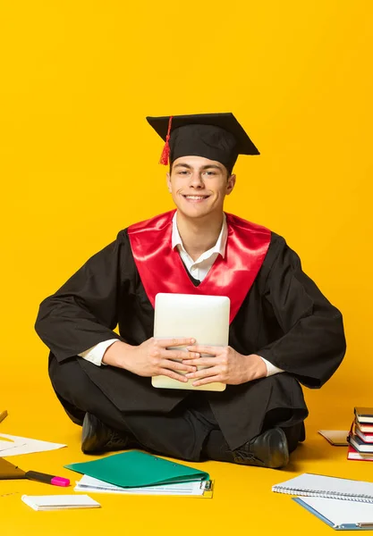 Retrato de un joven, estudiante con gorra de graduación y vestido sentado en el suelo alrededor de libros aislados sobre fondo amarillo del estudio —  Fotos de Stock