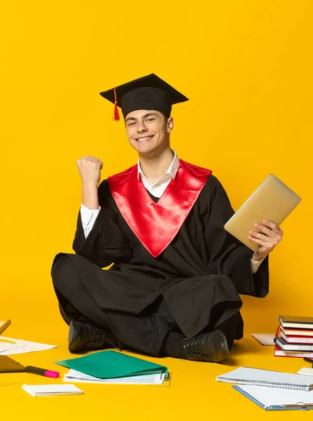 Retrato de un joven, estudiante con gorra de graduación y vestido sentado en el suelo alrededor de libros aislados sobre fondo amarillo del estudio. Exitoso. —  Fotos de Stock