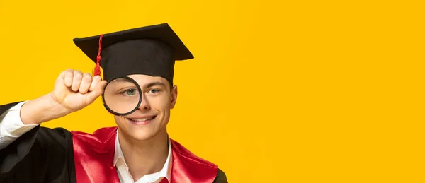 Retrato del joven sonriente, estudiante con gorra de graduación y vestido mirando con lupa aislada sobre fondo amarillo del estudio —  Fotos de Stock