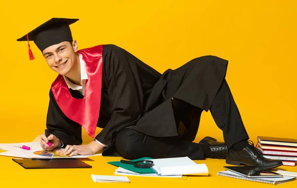 Retrato del joven sonriente, estudiante con gorra de graduación y vestido acostado en el suelo y haciendo tareas aisladas sobre fondo amarillo del estudio —  Fotos de Stock