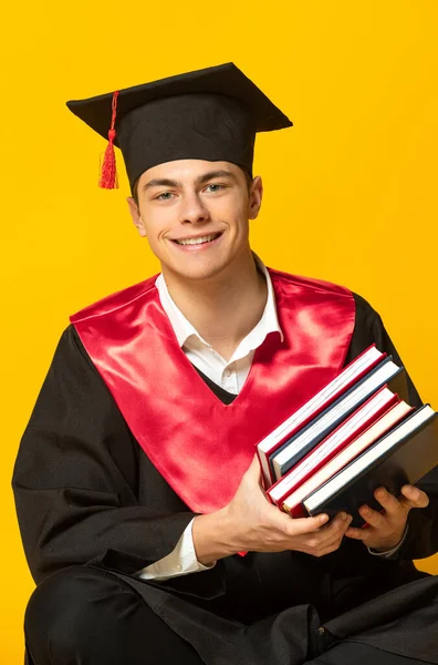 Retrato del joven sonriente, estudiante con gorra de graduación y vestido con libros posando aislados sobre fondo amarillo del estudio —  Fotos de Stock