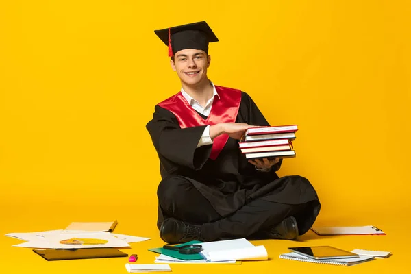 Retrato del joven sonriente, estudiante con gorra de graduación y vestido con libros sentados en el suelo aislados sobre fondo amarillo del estudio —  Fotos de Stock