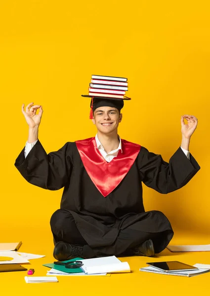 Retrato de un joven sonriente, estudiante con gorra de graduación y vestido sentado en el suelo con libros en la cabeza aislado sobre fondo amarillo estudio —  Fotos de Stock