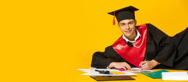 Retrato de un hombre joven con gorra de graduación y vestido acostado en el suelo artículos escolares aislados sobre fondo de estudio amarillo —  Fotos de Stock