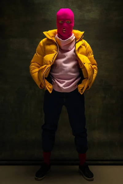 Studio di giovane anonimo in passamontagna rosa e piumino giallo, cappotto isolato su sfondo vintage scuro. Concetto di sicurezza, arte, moda — Foto Stock