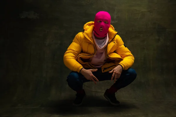 工作室拍摄的年轻的无名男子穿着粉红色的巴拉克拉瓦和黄色的羽绒服，外套孤立在黑暗的复古背景。安全、艺术、时尚的概念 — 图库照片