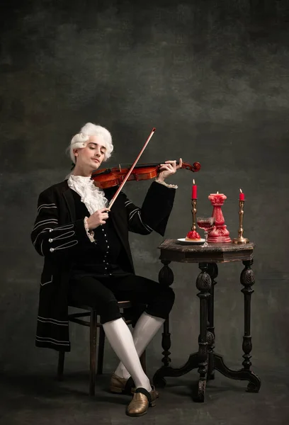 Portrait of young man wearing wig and vintage medieval outfit like famous composer playing violin isolated on dark green vintage background. — Stock Photo, Image