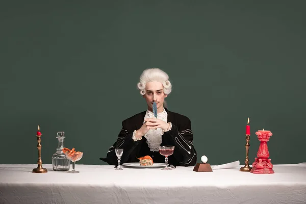 Arckép fiatal elegáns férfi peruke és vintage kabát ül asztalnál elszigetelt sötétzöld háttér. Retro stílus, a korszakok fogalmának összehasonlítása. — Stock Fotó