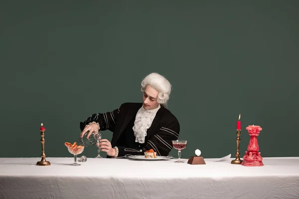 Arckép fiatal elegáns férfi peruke és vintage kabát ül asztalnál elszigetelt sötétzöld háttér. Retro stílus, a korszakok fogalmának összehasonlítása. — Stock Fotó
