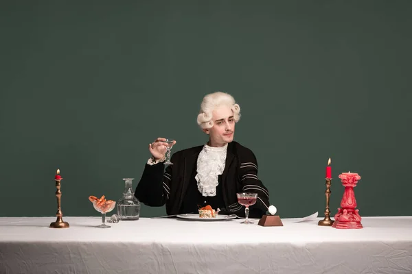 Porträt eines jungen eleganten Mannes in Peruke und Vintage-Jacke, der isoliert am Tisch auf dunkelgrünem Hintergrund sitzt. Retro-Stil, Vergleich des Epochenkonzepts. — Stockfoto