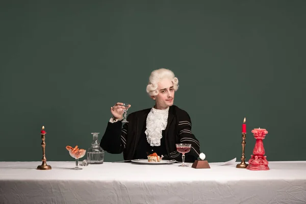 Portrait of young elegant man in peruke and vintage jacket sitting at table isolated on dark green background. Retro style, comparison of eras concept. — Stock Photo, Image