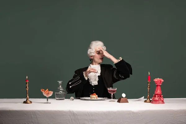 Portrét mladého elegantního muže v peruke a vintage bundě sedí u stolu izolované na tmavozeleném pozadí. Retro styl, srovnání konceptu éry. — Stock fotografie