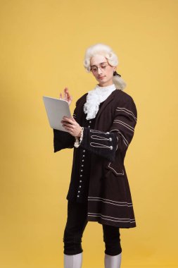 Sarı arka planda izole edilmiş beyaz peruklu ve klasik ortaçağ kıyafeti giymiş zarif genç bir adamın portresi. Sanat, güzellik, moda.
