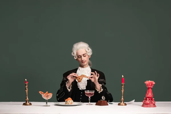 Portrait of young elegant man in peruke and vintage jacket sitting at table isolated on dark green background. Retro style, comparison of eras concept. — Stock Photo, Image