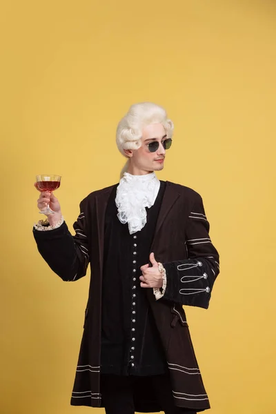 Retrato de hombre joven y elegante en peluca blanca y traje medieval vintage posando aislado sobre fondo amarillo. Arte, belleza, moda — Foto de Stock