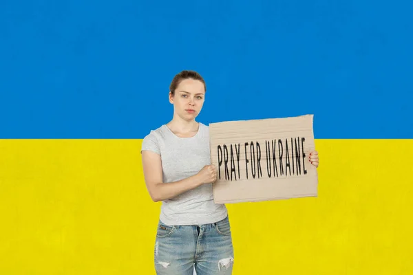 Conceptueel beeld. jong meisje holding tablet met belettering bidden voor Oekraïne geïsoleerd over Oekraïense blauwe en gele vlag achtergrond — Stockfoto