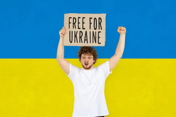 Konzeptionelles Bild. Junger Mann hält Tablette mit Schriftzug Free Ukraine isoliert über ukrainischer blau-gelber Flagge Hintergrund — Stockfoto