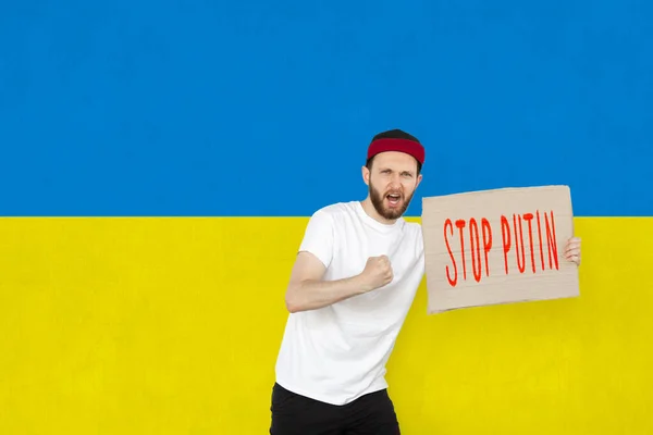 Konzeptionelles Bild. Junger Mann schreit, hält Tablet mit Schriftzug Stop Putin isoliert über ukrainischer blau-gelber Flagge Hintergrund — Stockfoto