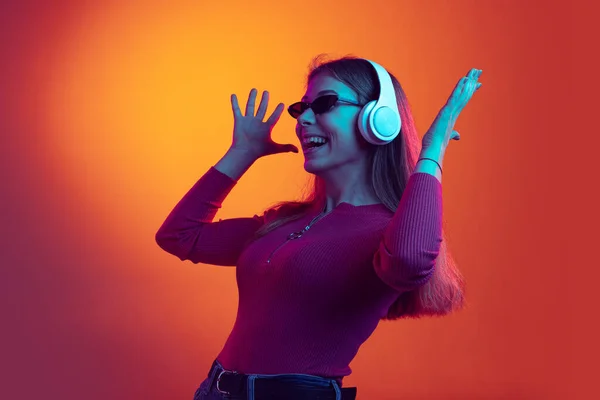 Retrato de joven hermosa chica feliz en auriculares escuchando música aislada sobre fondo naranja en luz de neón, filtro. Concepto de emociones, música —  Fotos de Stock