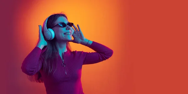 Leták. Portrét mladé krásné šťastné dívky ve sluchátkách poslech hudby izolované na oranžovém pozadí v neonovém světle, filtr. Pojetí emocí, hudby — Stock fotografie